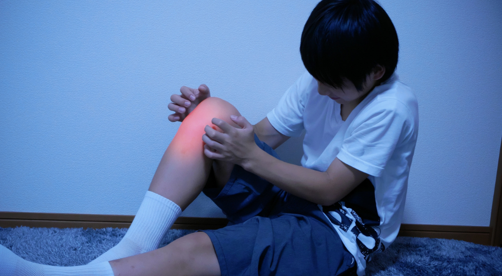 薄暗い部屋で床に座り込んだ小学生くらいの男の子が、【膝関節(脛骨粗面付近)】を手で押さえて患部が赤くハイライトされ、夜間の成長痛(オスグッド病など)やスポーツ後の痛みで辛そうにしている様子。