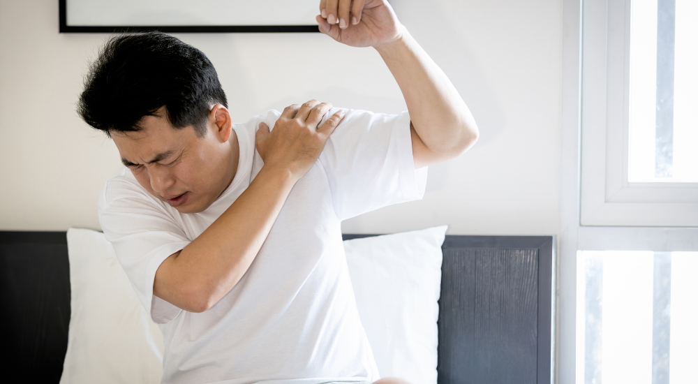 白いTシャツの男性が腕を上げようとした際、【肩関節(回旋腱板・三角筋周辺)】に激痛が走り、四十肩・五十肩(肩関節周囲炎、方関節拘縮)などで苦しそうに顔をしかめている様子。