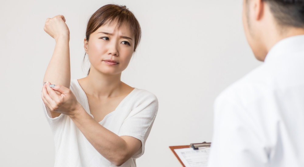 【肘関節】や【前腕】付近の痛みを訴え、不安そうな表情で専門家に相談している状況です。テニス肘などの腕の不調や、日常生活での辛さをカウンセリングで伝えている場面です。