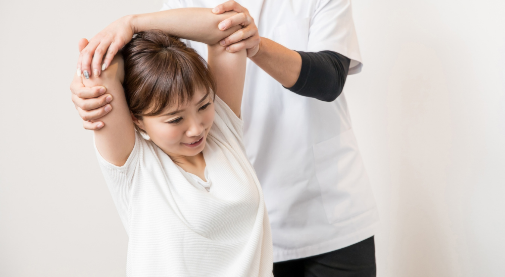 座った状態の女性患者に対し、施術者が腕を後ろに引いて【肩関節】や【大胸筋】を大きくストレッチしている状況。胸が開いて呼吸が深くなり、爽快な気分で笑顔になっている感情。